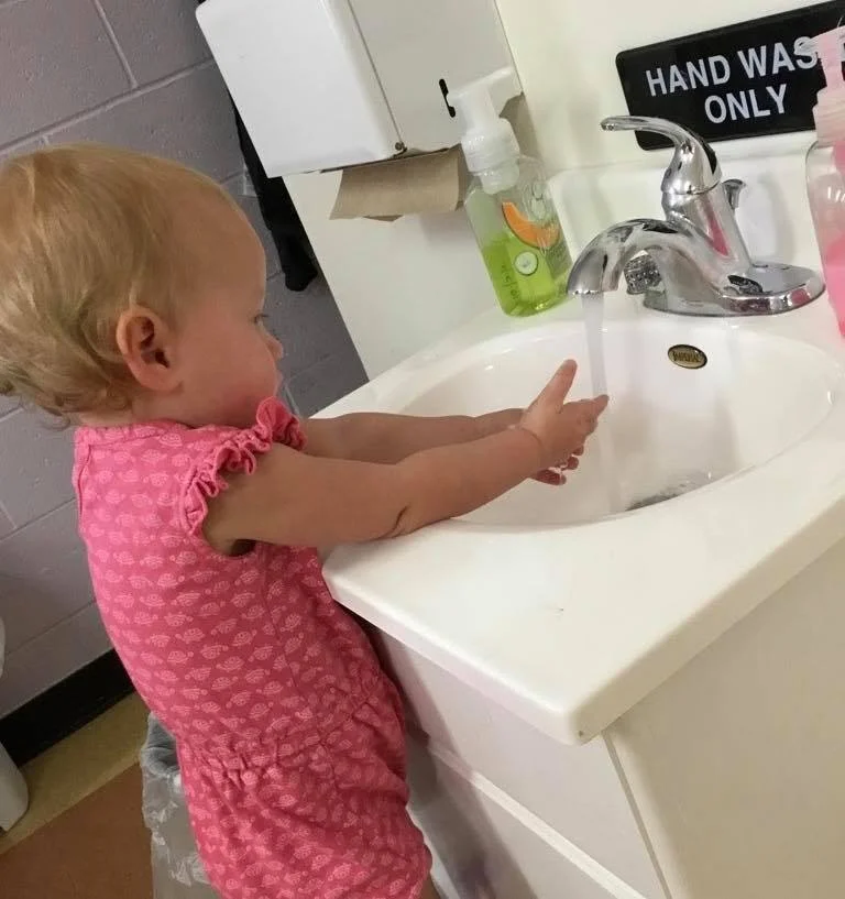 Toddler in a pink dress standing at a white sink, washing hands under running water with soap nearby.