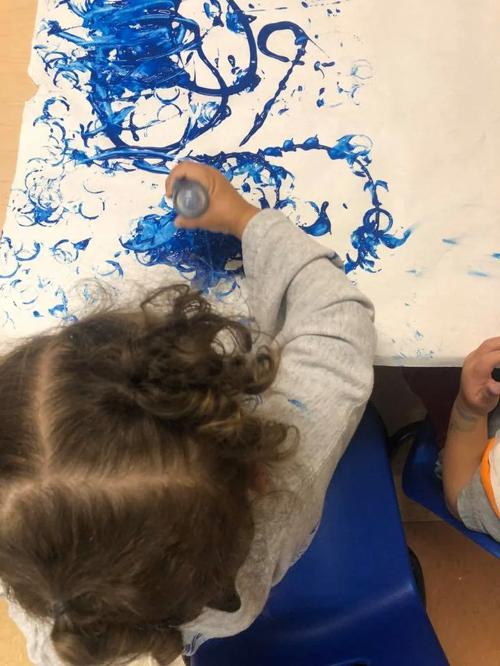 Child's hand presses blue paint onto a white sheet for finger painting, viewed from above; messy swirls of blue on paper and a gray sleeve nearby
