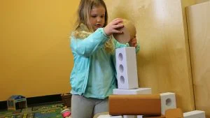Young girl stacking large wooden blocks and a cardboard tower during play in a classroom or playroom.