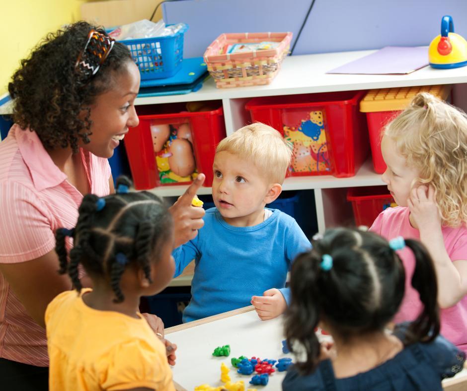 Teacher engages with four preschoolers at a table in a bright classroom, guiding a hands-on activity with small colorful pieces on the table.