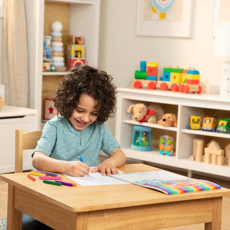 A smiling child with curly hair draws with a blue marker at a wooden table in a colorful playroom.