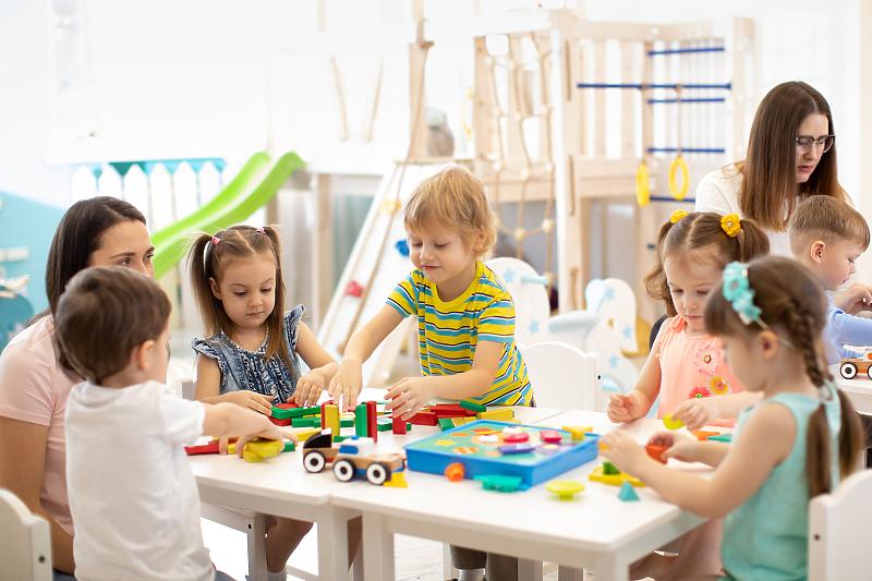 Children and caregivers play with colorful blocks at a bright classroom table, building together in a daycare setting.