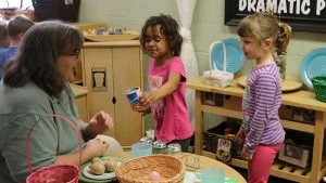 Teacher guiding two young girls in a classroom pretend‑play kitchen, pouring drinks at a table with dishes and baskets on display.