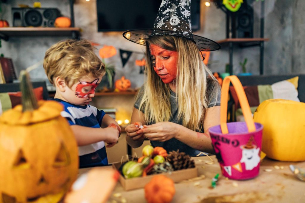 A woman wearing a witch hat helps a child with Halloween crafts at a festive table with pumpkins in the foreground.