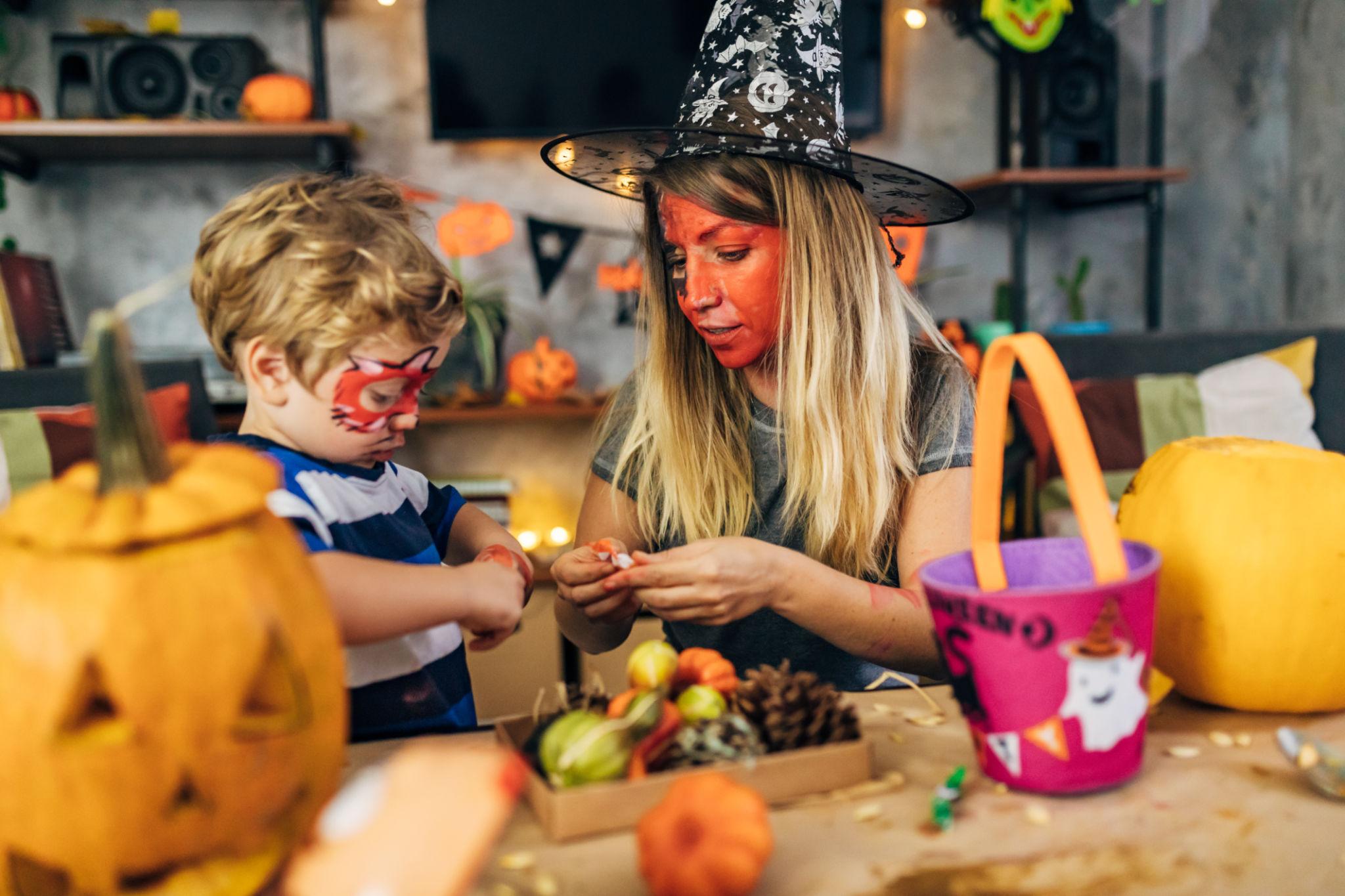 A woman wearing a witch hat helps a child with Halloween crafts at a festive table with pumpkins in the foreground.