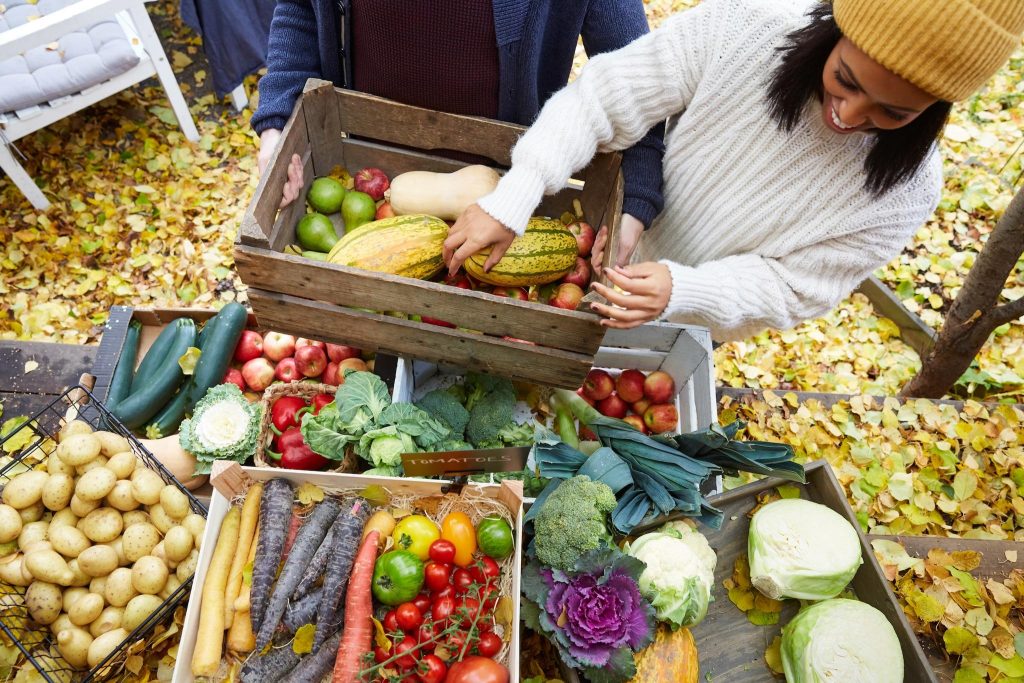 Two people in sweaters holding a wooden crate filled with autumn produce—pumpkins, squash, apples, and pears—at an outdoor market on fall leaves.