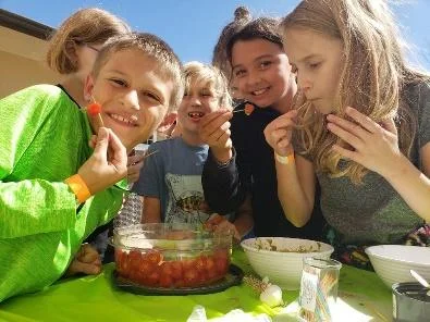 Five smiling children sit around a sunny outdoor table, sharing a bowl of cherries dessert.
