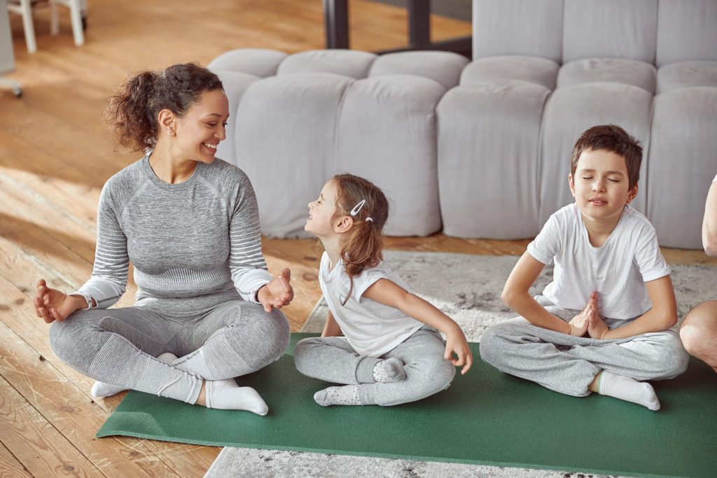 Woman and two children sit cross-legged on a green yoga mat, meditating together in a cozy living room.