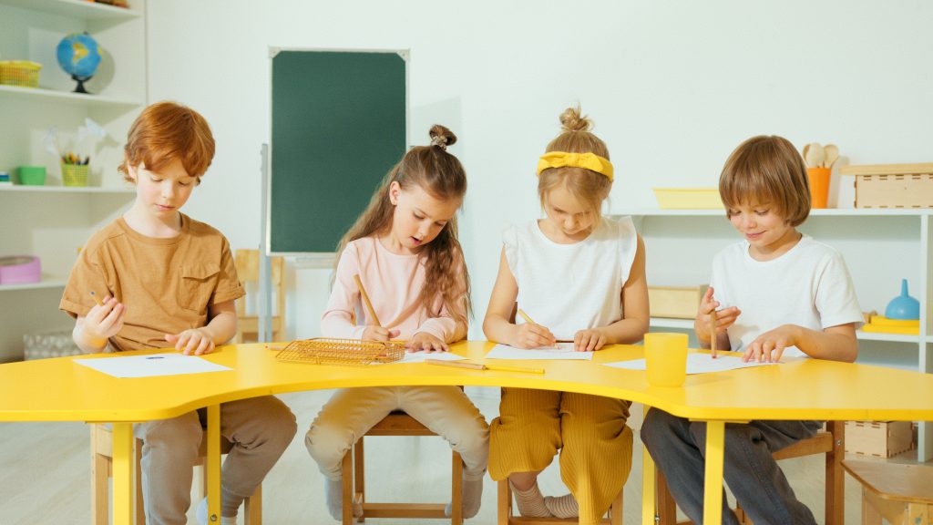Four kids sit at a bright yellow table, drawing with pencils in a classroom setting.